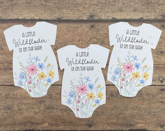 Könnte beinhalten: Drei weiße Papier-Babybodys mit einem Blumendesign und dem Text "A Little Wildflower is on the way!" darauf gedruckt.