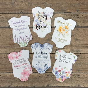 K&ouml;nnte beinhalten: F&uuml;nf wei&szlig;e Baby-Strampler-f&ouml;rmige Dankeskarten mit Blumendesigns und Text. Der Text enth&auml;lt "Baby in Bloom", "Vielen Dank f&uuml;rs Feiern mit uns!" und "Unser Baby bl&uuml;ht!" mit Daten und Namen.