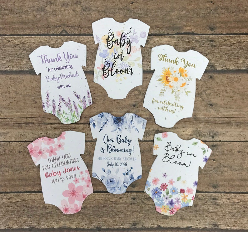 K&ouml;nnte beinhalten: F&uuml;nf wei&szlig;e Baby-Strampler-f&ouml;rmige Dankeskarten mit Blumendesigns und Text. Der Text enth&auml;lt "Baby in Bloom", "Vielen Dank f&uuml;rs Feiern mit uns!" und "Unser Baby bl&uuml;ht!" mit Daten und Namen.
