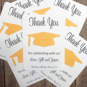 May include: Thank you cards with a graduation cap design. The cards are white with black text and a gold paper graduation cap. The text on the cards reads "Thank You" and "for celebrating with us! Love, Gill and Jason". The cards also include the text "Plant this in celebration. Class of 2029".