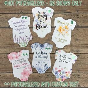 K&ouml;nnte beinhalten: F&uuml;nf wei&szlig;e Baby-Strampler-f&ouml;rmige Dankeskarten mit Blumendesigns. Der Text auf den Karten lautet "Vielen Dank f&uuml;r die Feier von Baby Michael mit uns!" "Vielen Dank f&uuml;r die Feier mit uns!" "Vielen Dank f&uuml;r die Feier von Baby Jones 12. Mai 2028" "Unser Baby bl&uuml;ht! Baby Shower von Melissa 10. Juli 2029" und "Baby in Bloom!"