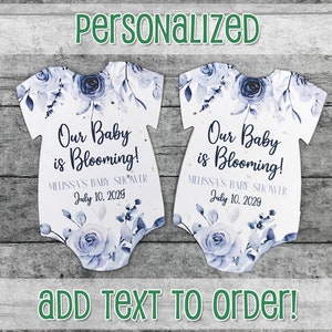 K&ouml;nnte beinhalten: Zwei wei&szlig;e, babyf&ouml;rmige Saatpapier-Geschenk-Anh&auml;nger mit blauen Aquarell-Blumenmustern. Der Text lautet "Unser Baby bl&uuml;ht!" und "Melissas Babyparty 10. Juli 2029".