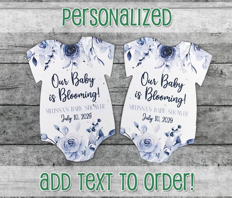 K&ouml;nnte beinhalten: Zwei wei&szlig;e, babyf&ouml;rmige Saatpapier-Geschenk-Anh&auml;nger mit blauen Aquarell-Blumenmustern. Der Text lautet "Unser Baby bl&uuml;ht!" und "Melissas Babyparty 10. Juli 2029".
