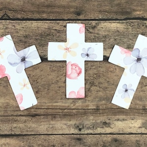 May include: Three wooden crosses with a floral pattern. The crosses are white with pink, purple, and yellow flowers.