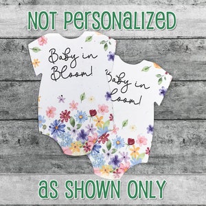 K&ouml;nnte beinhalten: Zwei wei&szlig;e Baby-Bodys mit einem Blumenmuster und dem Text "Baby in Bloom!" in schwarzem Stift.