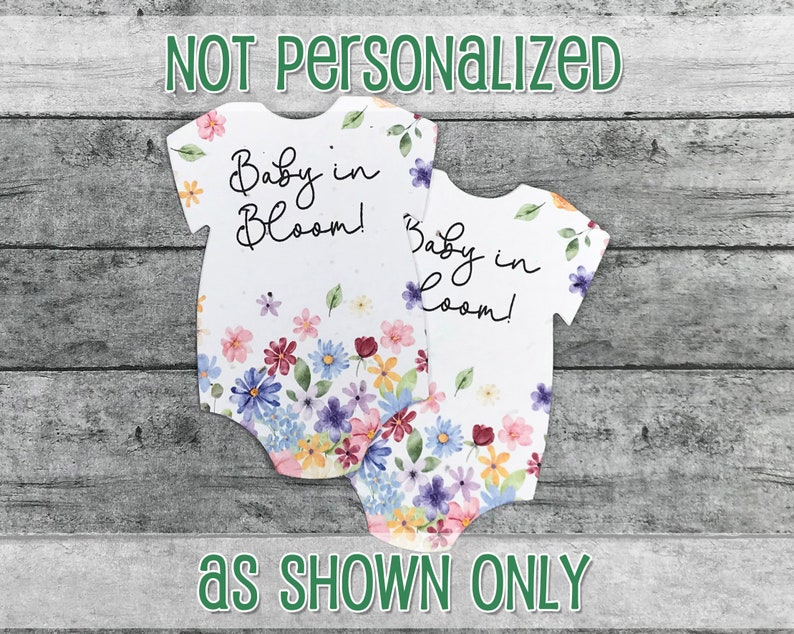 K&ouml;nnte beinhalten: Zwei wei&szlig;e Baby-Bodys mit einem Blumenmuster und dem Text "Baby in Bloom!" in schwarzem Stift.