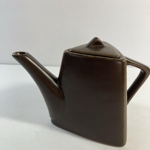 May include: A brown ceramic teapot with a geometric design. The teapot has a square body, a triangular lid, and a curved spout. The handle is a simple, angular loop. The teapot is a solid, dark brown color.