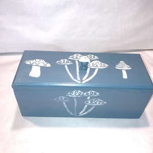 May include: A rectangular blue wooden box with a lid featuring a small white knob. The top and front surfaces are decorated with white stenciled mushroom illustrations, including various cap shapes and stem designs painted in a matte white finish.