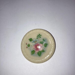 May include: A round, off-white decorative coaster with a floral design. The coaster features a central arrangement of pink, white, and blue flowers with green leaves. The edge has a rope-like texture.