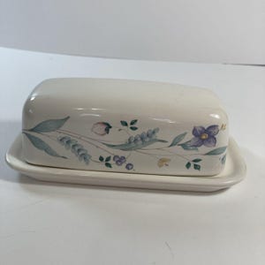 May include: Cream-colored ceramic butter dish with a lid. The dish and lid are decorated with a floral pattern in shades of purple, blue, and green. The butter dish is rectangular with rounded edges.