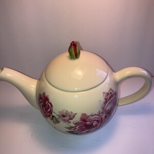 May include: Cream-colored ceramic teapot with a floral rose design in shades of pink and purple. The lid is topped with a green and red bud-shaped handle. Features a curved spout and handle.