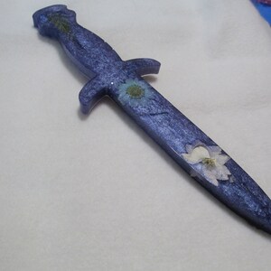 May include: A purple dagger with embedded dried flowers and glitter. The dagger has a silver-colored handle and a pointed tip.
