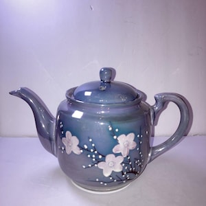 May include: A ceramic teapot with a lid, spout, and handle. The teapot is a gradient of blue and gray with white floral designs. The teapot sits on a white base and is suitable for serving tea.