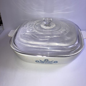 May include: Square, off-white casserole dish with a clear glass lid. The dish has a floral pattern in blue and two small handles. The lid has a clear knob handle.