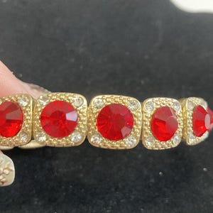 May include: Gold-toned bracelet featuring square links with large, round, red gemstones. Each link is framed with smaller, clear crystals. The bracelet is on a stretchy band, and the gemstones are a vibrant red color.