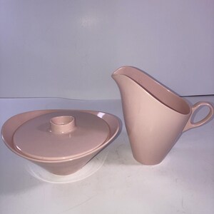 May include: A light pink ceramic creamer and a matching lidded sugar bowl. The creamer features an angled rim and a small handle. The sugar bowl has a circular lid with a central knob. Both pieces have a smooth, solid-colored finish.