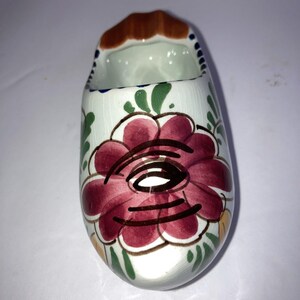 Può includere: Un vaso a forma di zoccolo in ceramica, dipinto di bianco con un grande motivo floreale bordeaux. Lo zoccolo ha una parte superiore marrone e accenti di foglie verdi. L'interno è bianco e il design ricorda l'arte popolare olandese.