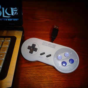 May include: A gray Super Nintendo controller with a USB connector. The controller has a D-pad, six buttons, and the text "SUPER NINTENDO" printed on it.