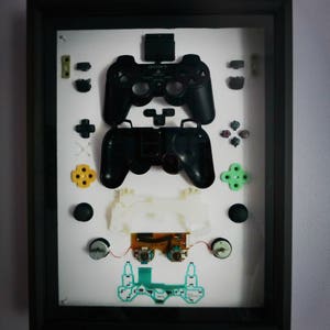 May include: A framed display of disassembled parts of a black Sony PlayStation controller. The display includes the controller's face buttons, analog sticks, and other components.