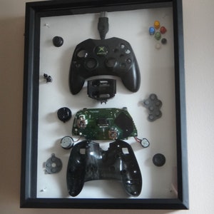 May include: A disassembled Xbox controller in a black frame. The controller is separated into its individual components, including the face buttons, the circuit board, and the shell. The controller is displayed on a white background.