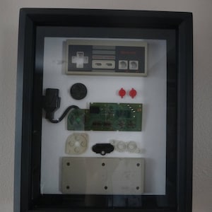 May include: A framed display of disassembled parts from a vintage Nintendo Entertainment System (NES) controller. The display includes the controller faceplate, buttons, a circuit board, and a connector cable. The parts are arranged on a white background within a black frame.