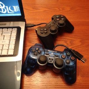 May include: Two black and blue Sony PlayStation 2 controllers connected to a laptop via USB cables. The controllers have buttons, joysticks, and a start button.