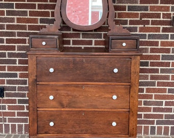 May include: A dark brown wooden dresser with five drawers and a beveled mirror on top. The dresser has white porcelain knobs on the drawers and a scalloped base.