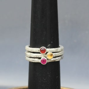 Op de afbeelding: Stapelbare sterling zilveren ringen met getextureerde banden, elk met een andere edelsteen: rood, geel en roze. De ringen worden gepresenteerd op een zwarte ringhouder tegen een grijze achtergrond.