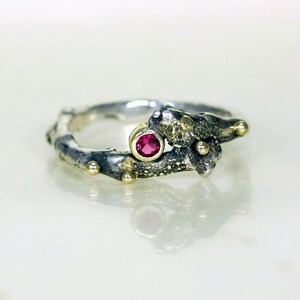 May include: A silver ring with a textured, branch-like design. The ring has a small, round, red gemstone set in a gold setting.