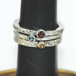 May include: Three silver stacking rings with a hammered finish, each featuring a different coloured gemstone: red, blue, and orange.