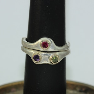 Op de afbeelding: Twee zilveren ringen met een getextureerde afwerking. De bovenste ring heeft een rode edelsteen, en de onderste ring heeft een paarse en een groene edelsteen.