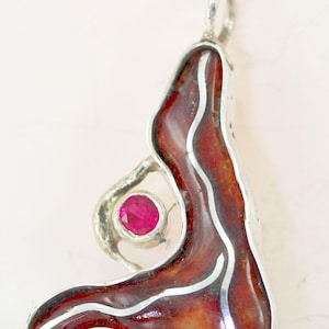 Puede incluir: Colgante de plata con un diseño abstracto. Presenta un esmalte marrón rojizo con detalles blancos y una gema rosa redonda. El colgante está diseñado con un lazo en la parte superior para una cadena.