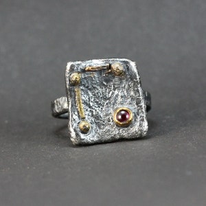 Op de afbeelding: Een zilveren ring met een getextureerd, rechthoekig ontwerp. De ring heeft een kleine, rode edelsteen in het midden en gouden accenten.