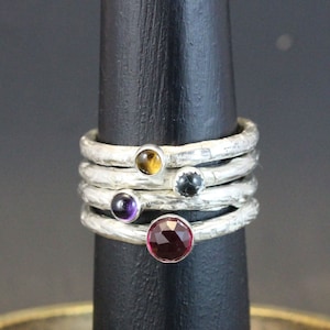 Op de afbeelding: Vijf zilveren ringen met verschillende gekleurde edelstenen. De ringen zijn op elkaar gestapeld. De edelstenen zijn geelbruin, zwart, paars en rood.