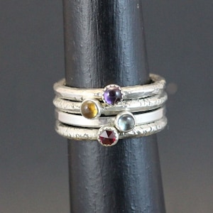 Op de afbeelding: Vier zilveren stapelringen met getextureerde banden en kleine edelstenen. De edelstenen zijn amethyst, topaas, aquamarijn en granaat.