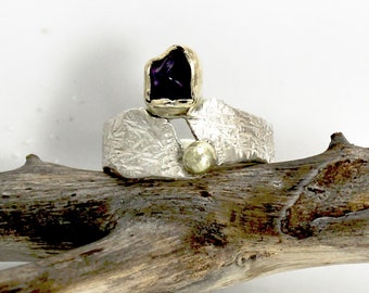 May include: A silver ring with a textured band and a purple amethyst gemstone set in gold. A small gold bead is also set in the band.