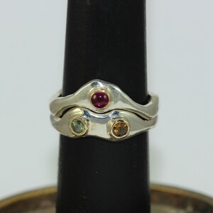 Op de afbeelding: Twee zilveren ringen met een golvend ontwerp. De bovenste ring heeft een enkele rode edelsteen, en de onderste ring heeft twee edelstenen, een groene en een oranje.