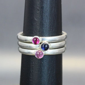 Op de afbeelding: Drie zilveren ringen met edelstenen. De bovenste ring heeft een roze edelsteen, de middelste ring heeft een blauwe edelsteen en de onderste ring heeft een roze edelsteen.