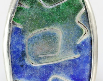 Puede incluir: Colgante ovalado con marco plateado. El colgante presenta un diseño verde y azul con detalles de alambre plateado. Los colores se mezclan, creando un aspecto artístico único. El colgante es probablemente de esmalte.