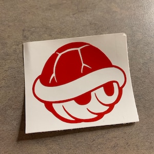 May include: A red cartoon turtle shell sticker on a white background. The shell has a smiling face and is a popular video game reference. The sticker is rectangular and sits on a gray surface.
