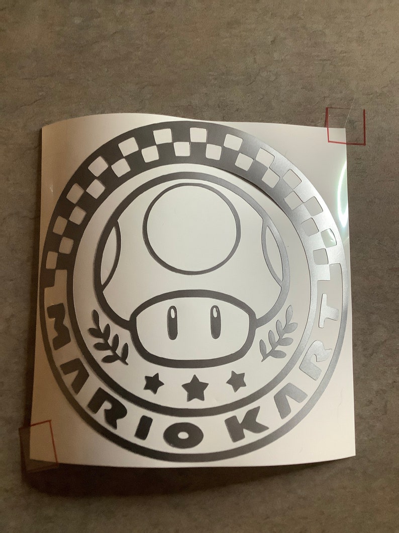May include: A silver vinyl sticker with a black and white chequered circle around a mushroom character with the text "Mario Kart" in a circle around the mushroom.