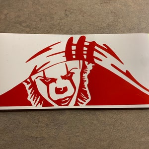 May include: Red vinyl decal of a clown's face peeking out from behind a red and white striped pattern. The clown has a menacing expression with a wide smile and sharp teeth.