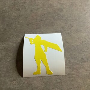 May include: A yellow silhouette of a character holding a large sword, on a white square sticker. The character is in a dynamic pose, suggesting action. The sticker is placed on a gray surface.