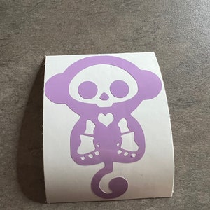 May include: A purple vinyl sticker of a cartoon monkey with a skull for a head and a heart on its chest.