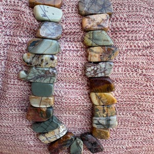 May include: A necklace composed of rectangular, polished stones in shades of brown, gray, green, and cream. The stones are strung together, creating a U-shaped design. The necklace is displayed on a textured, reddish-brown surface.