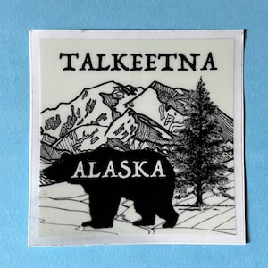 May include: A white sticker with a black silhouette of a bear and a pine tree. The text "TALKEETNA ALASKA" is printed in black on the sticker.