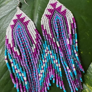 May include: A pair of long, beaded earrings with a geometric pattern in shades of white, purple, blue, and silver. The earrings are made with small, seed beads and have a fringe-like design.