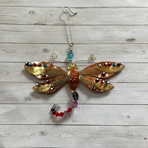 May include: A metal dragonfly ornament with a copper and gold finish. The dragonfly has red, green, and blue glass beads for accents. The ornament has a wire hanger.