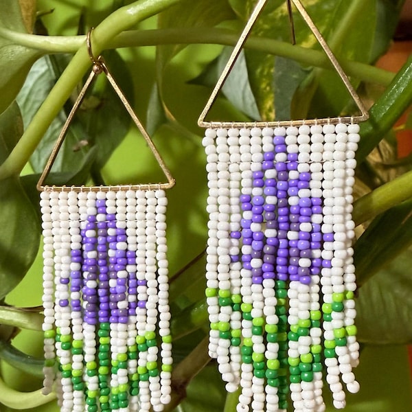 May include: A pair of dangle earrings featuring gold-tone triangular frames with white, purple, and green seed beads arranged in a floral pattern. The beads form a vertical flower design with a purple bloom and green stem against a white background.