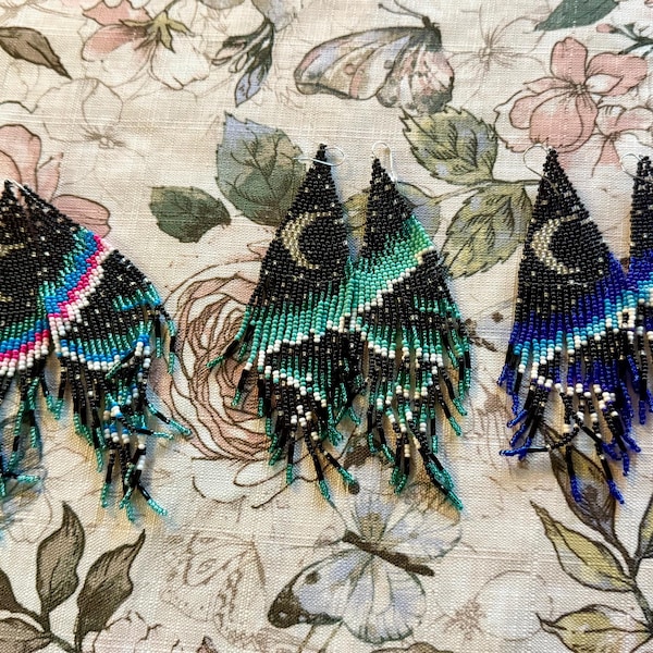 May include: Three pairs of beaded earrings with a crescent moon and star design. The earrings are black with blue, green, and white accents. The earrings are handmade and have a fringe design.